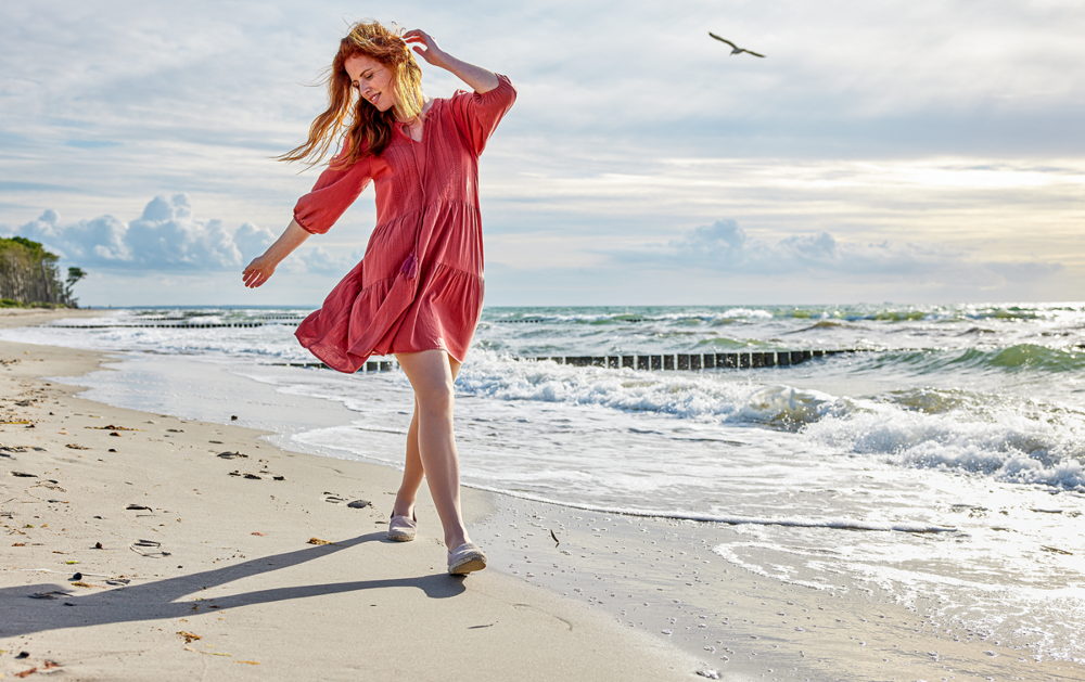 Vrouw in rode jurk loopt langs de kustlijn met therapeutische kousen aan, op een zonnige dag aan zee